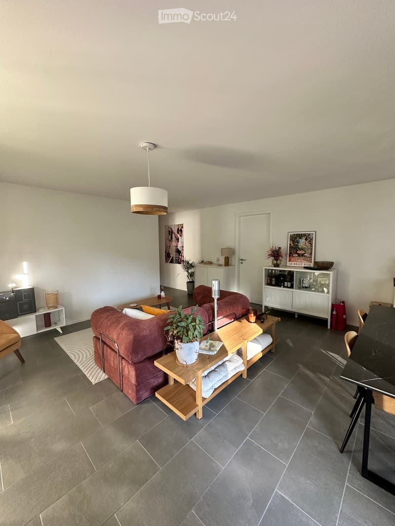 Modern living room with a red sofa, wooden coffee table, side tables, and a potted plant. The room features a grey tiled floor, white walls, and a hanging pendant light.