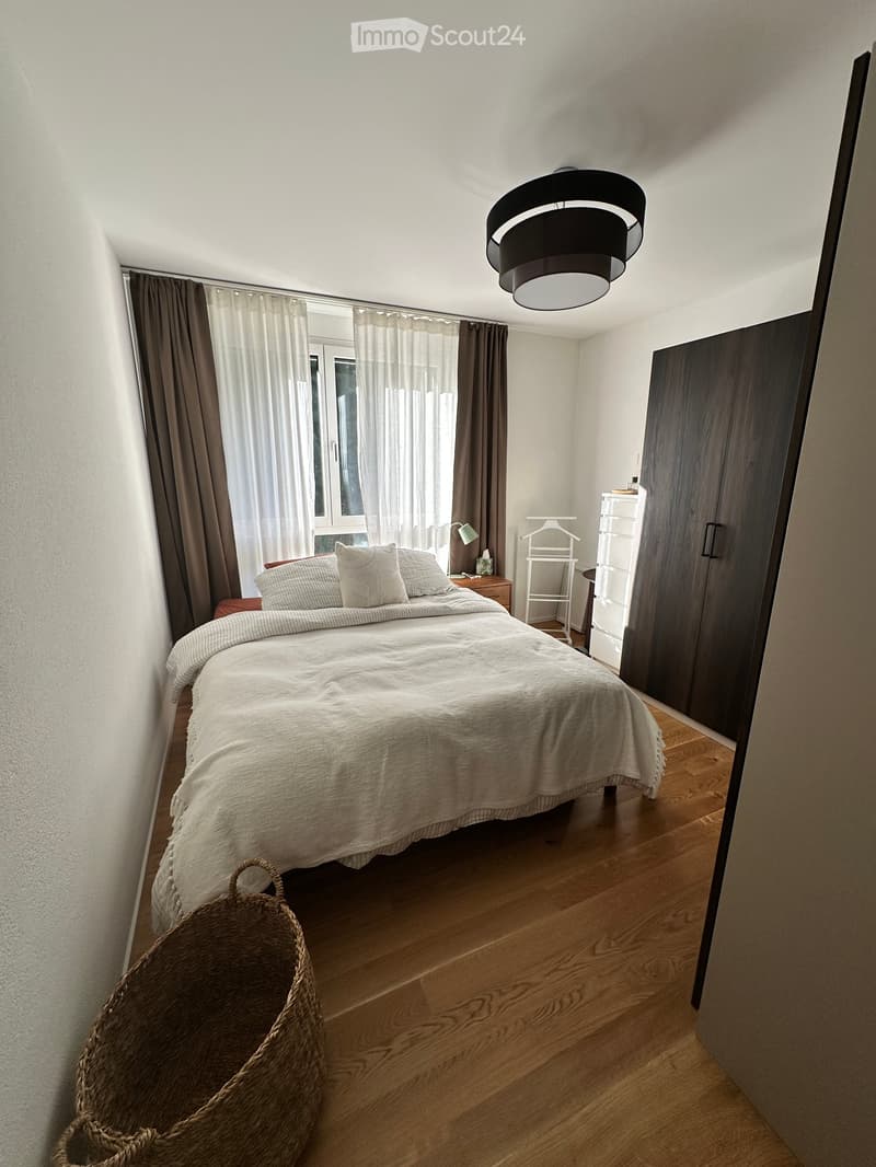 Cozy bedroom with a large bed, nightstand, lamp, wardrobe, and wicker basket. Light-colored bedding and curtains, wooden flooring.