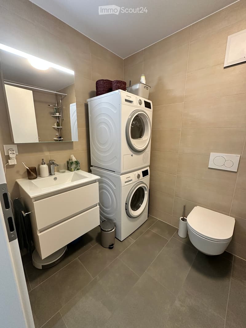 Modern bathroom with beige tiles, a white sink with a mirror cabinet, a toilet, and a stacked washing machine and dryer.