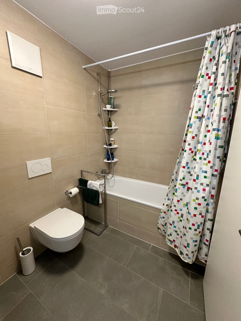 Modern bathroom with beige tiled walls, a white toilet, a bathtub with a colorful shower curtain, a towel rack with towels, and a shower shelf with toiletries.