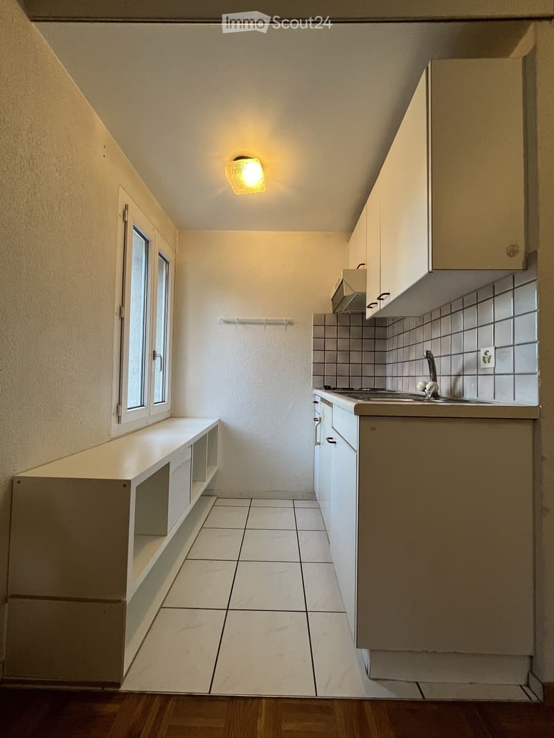 Empty kitchen with white tiled floor, white cabinets, and a window. The space includes a sink with a faucet and a built-in bench with storage compartments.