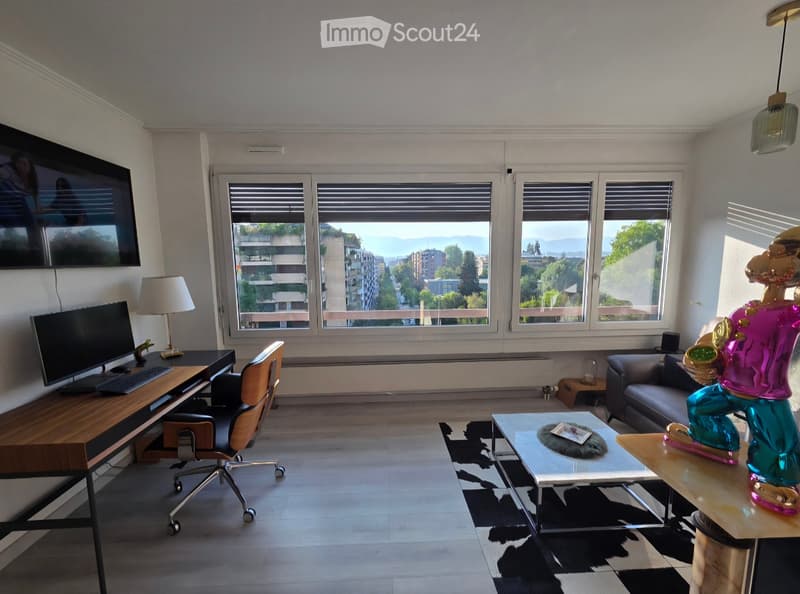 Modern living room with large windows offering a scenic view, featuring a sofa, coffee table, desk with a chair, and a decorative lamp.