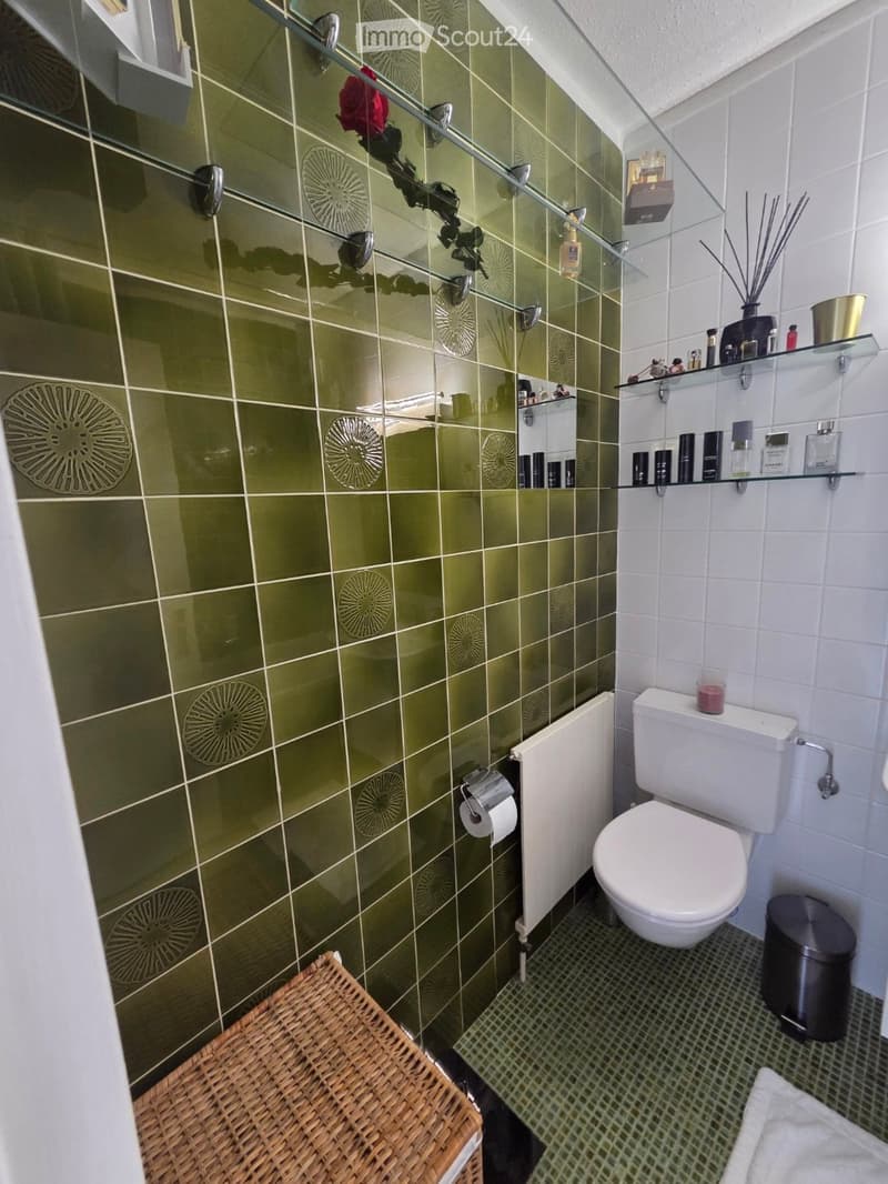 Modern bathroom with green tiled walls, white toilet, glass shelves holding perfume bottles and candles, wicker basket, radiator, and a trash bin.