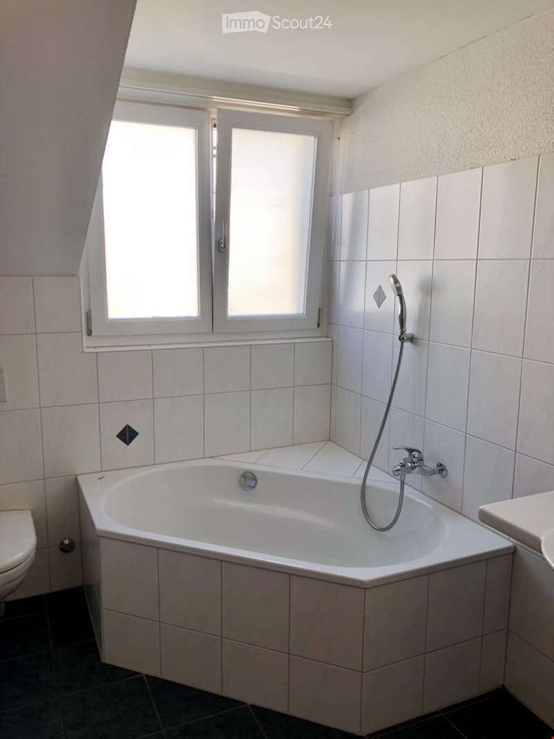 Modern bathroom with a corner bathtub, tiled walls, and a window above the tub. Includes a toilet and a showerhead attached to the wall.