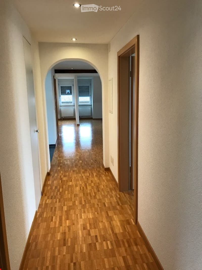 A narrow hallway with wooden flooring, white walls, and recessed lighting. An arched doorway leads to another room with a visible window and door.