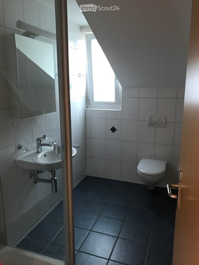 Modern bathroom with white tiles, a sink with a chrome faucet, a mirror, and a toilet. The room features a slanted ceiling and a small window allowing natural light.