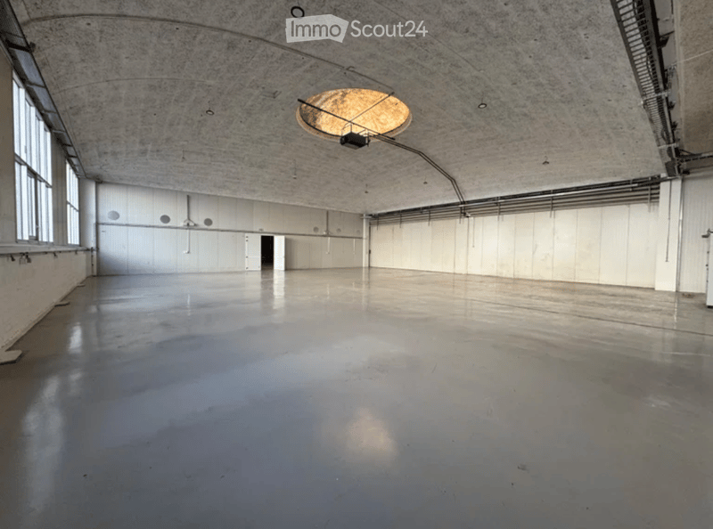 Large, empty industrial-style room with concrete floors and walls, large windows on one side, and a single circular light fixture on the ceiling.