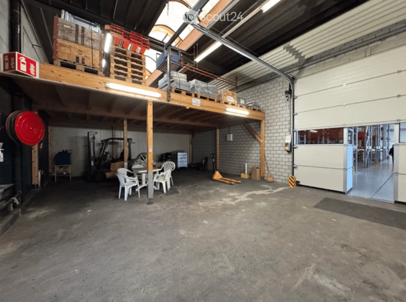 Large industrial-style warehouse with concrete floor, exposed wooden beams, and a raised platform with storage crates. Includes a forklift, a small seating area with chairs and a table, and a large garage door.