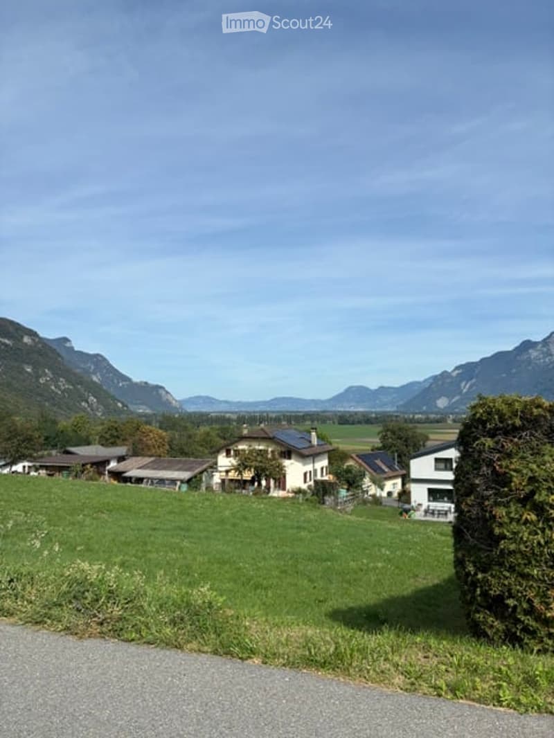 A peaceful rural landscape with a cluster of houses, green fields, and distant mountains under a clear blue sky.