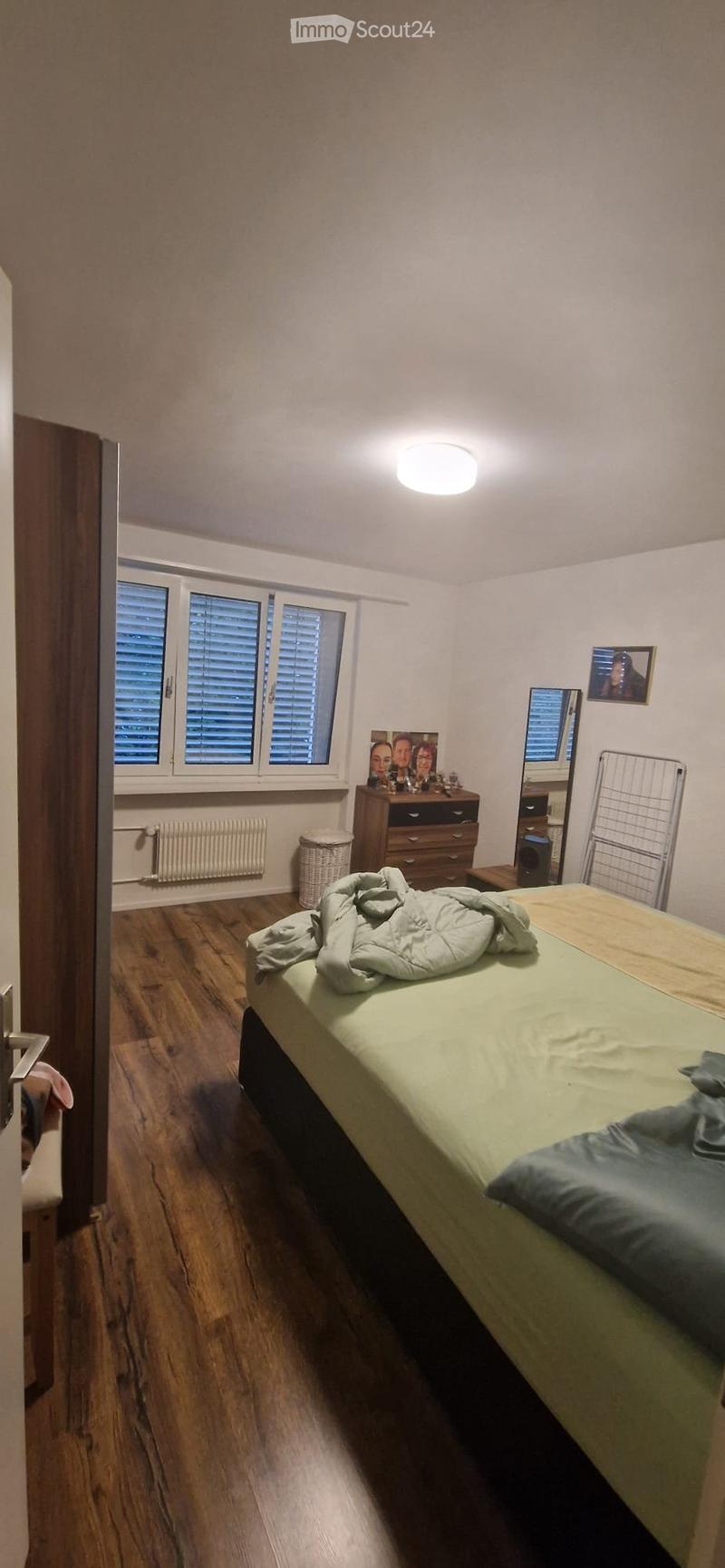 Bedroom with wooden flooring, large window with blinds, bed with green bedding, nightstand, mirror, radiator, picture frame, wicker basket, and speaker.