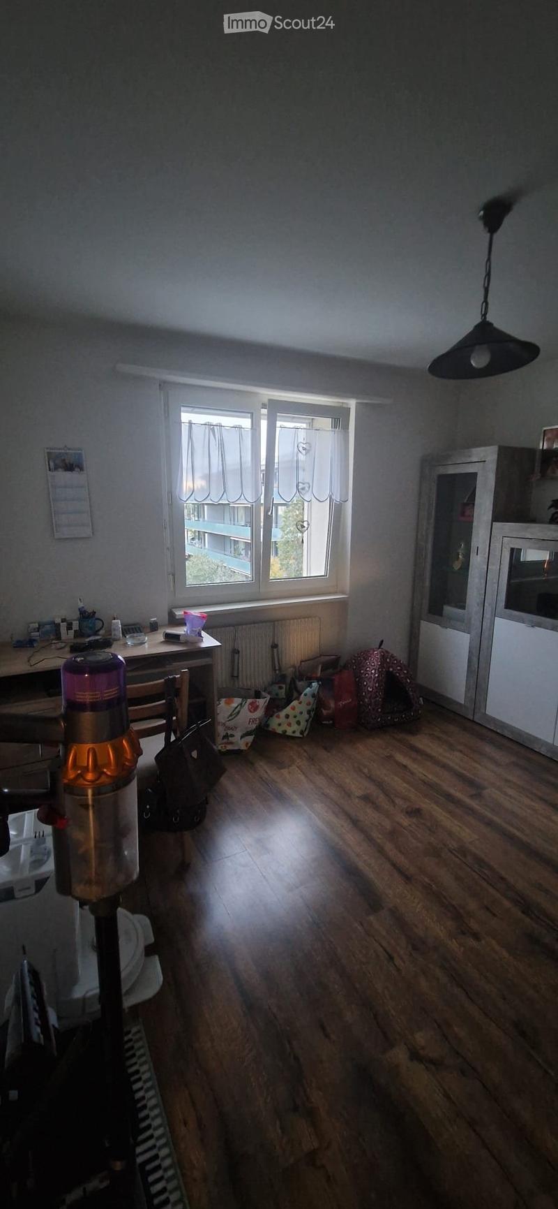 A furnished room with wooden flooring, a large window with white curtains, a table with a chair, a hanging lamp, a dog bed, and a small cabinet.