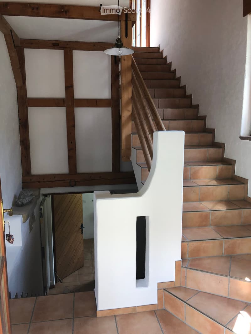 Spacious hallway with wooden staircase, white walls, and exposed wooden beams. Features a modern pendant light and a doorway leading to another room.