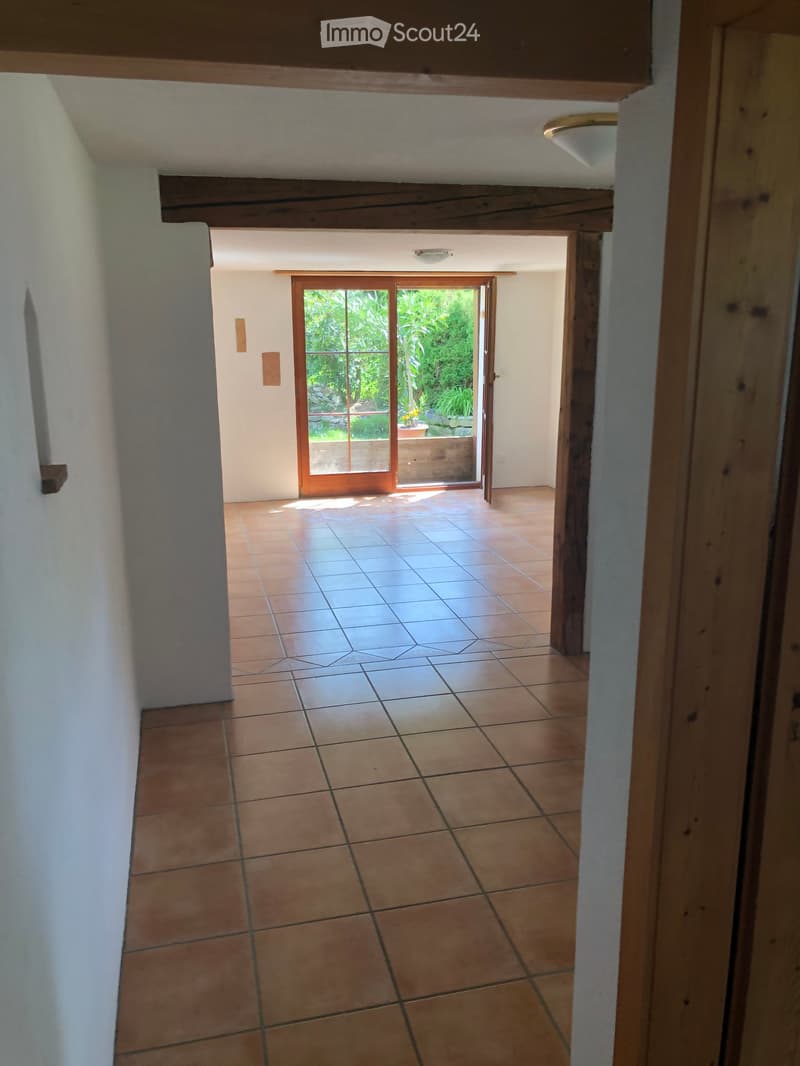 Empty hallway with tiled floor, white walls, wooden beams, and a sliding glass door leading to a garden area.