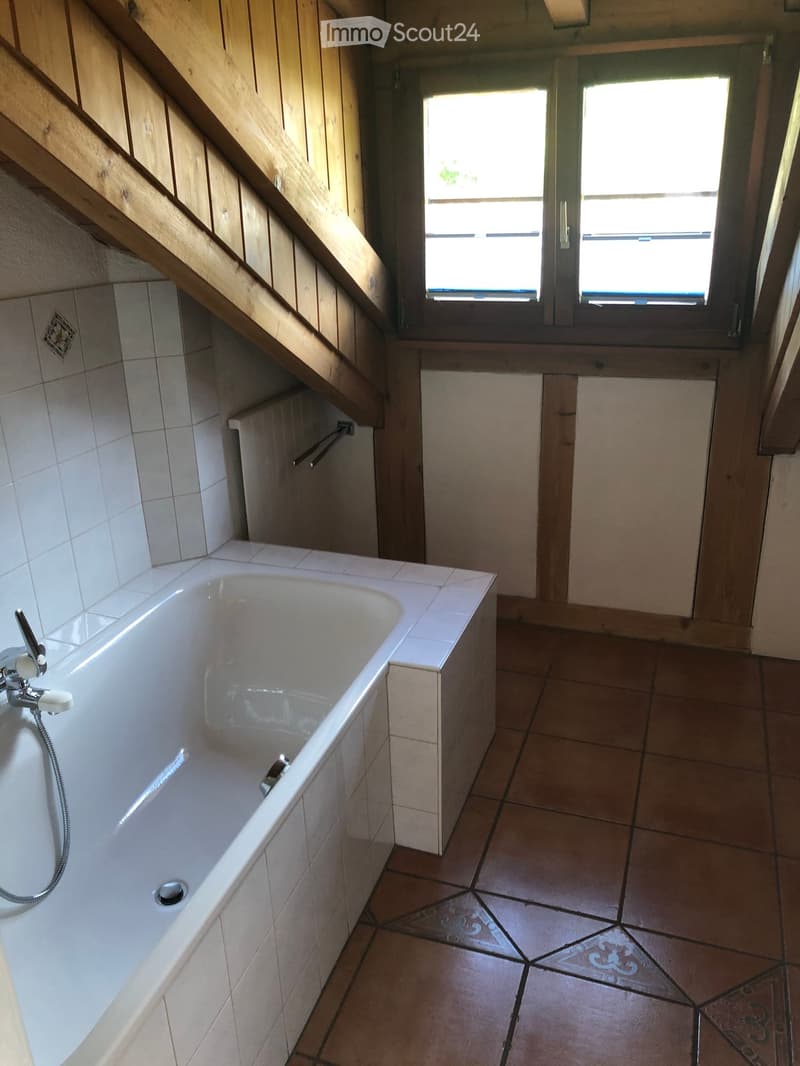 Bathroom with a white bathtub, tiled walls, and wooden flooring. Features a window with wooden frame and a small wooden shelf.