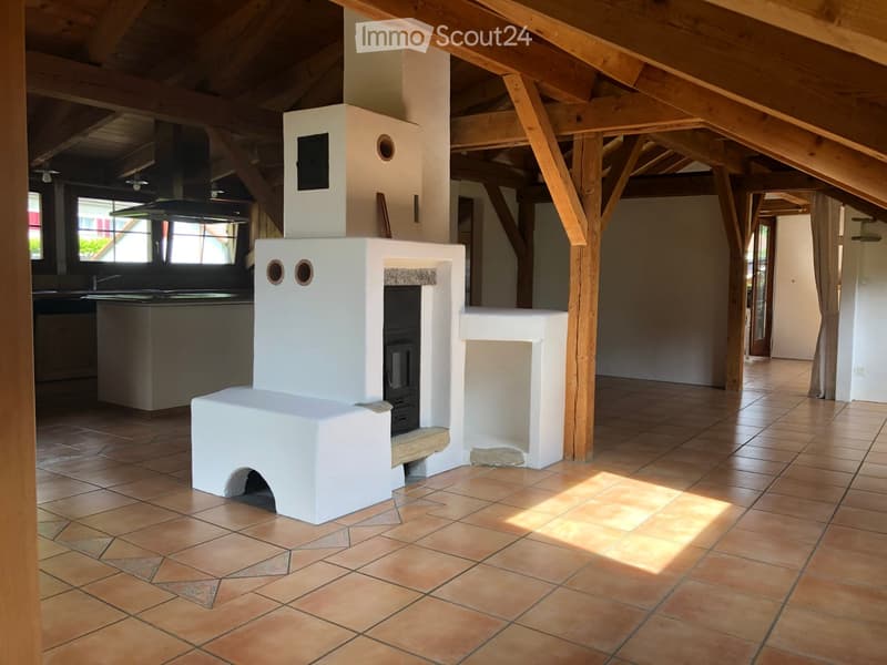 Open-plan living space with exposed wooden beams, a white fireplace, and a kitchen area visible in the background.