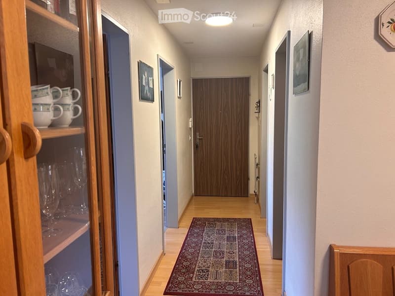 A narrow hallway with wooden flooring, a patterned rug, and several doors leading to different rooms. The walls are painted in a light color, and there are framed pictures on the walls. A wooden cabinet with glass doors is visible on the left side, contai