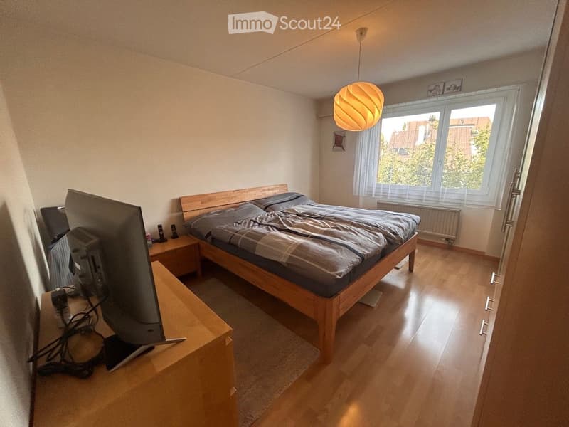 A well-lit bedroom with a wooden bed, nightstand, and a large window offering a view of greenery outside. The room features a modern lamp and a screen on a wooden stand.