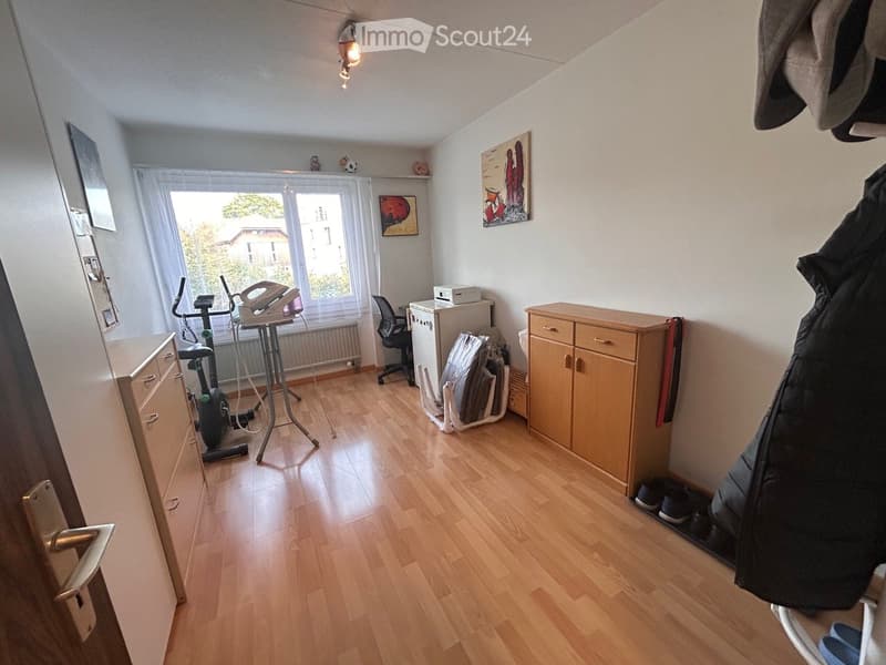 Well-lit room with wooden flooring, exercise bike, ironing board, chair, small refrigerator, cabinet, and artwork on the walls.
