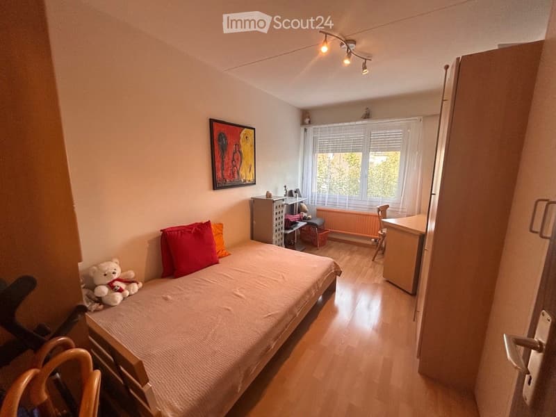 Cozy bedroom with a single bed, colorful pillows, a stuffed animal, a small table, and a window with blinds.