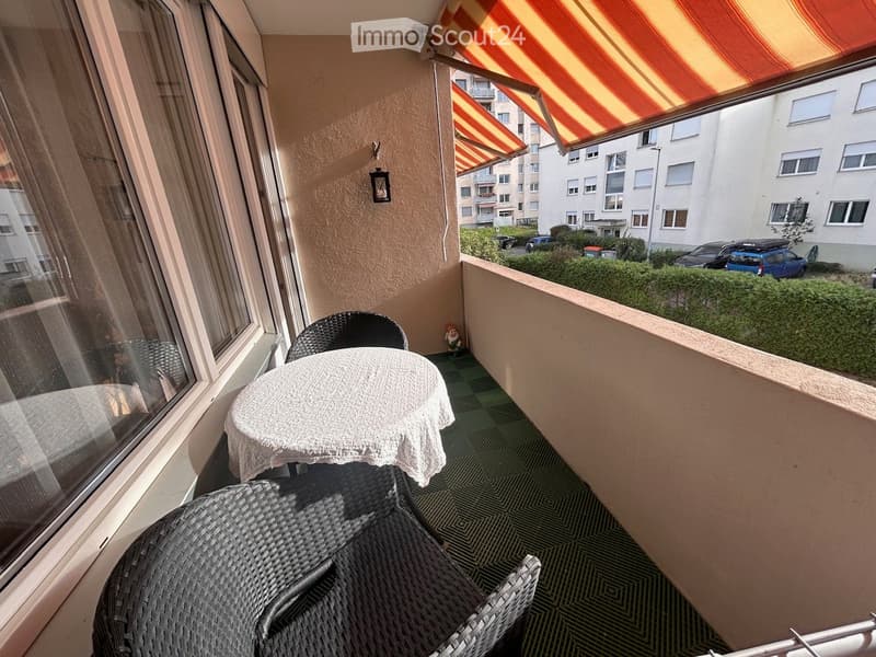 Balcony with a table and two chairs, a small decorative figure, and a retractable awning. The view shows a residential area with parked cars and buildings.