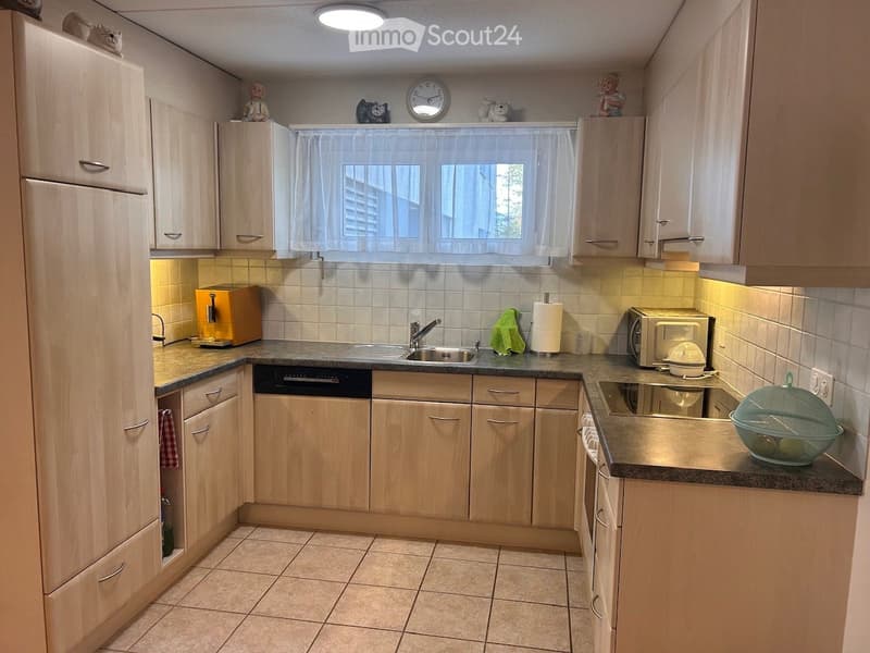 The kitchen features light wooden cabinets, a tiled backsplash, and a window with a white curtain. Appliances include a refrigerator, sink, dishwasher, microwave, toaster, and oven. A paper towel roll, dish rack, and bowl are on the counter.