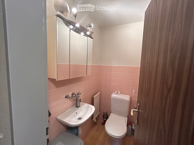 A small bathroom with pink tiles, a white sink, a toilet, a mirrored cabinet, and a wooden floor.