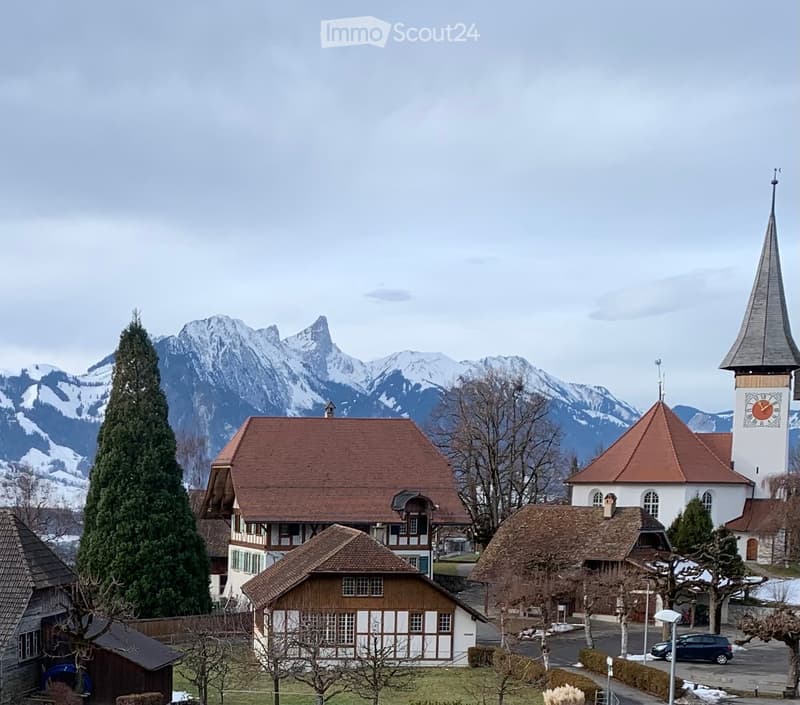 Village scene with traditional Swiss houses, a church with a tall spire, and snow-capped mountains in the background.