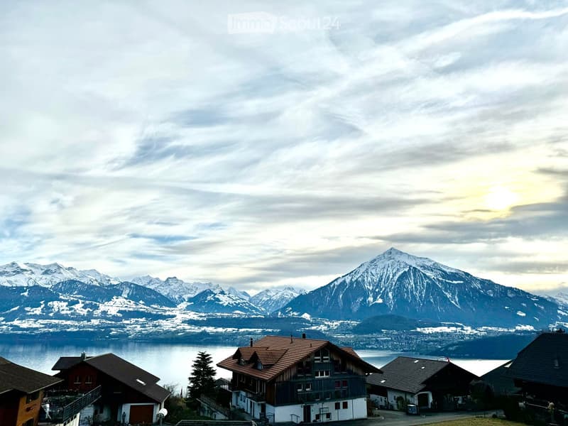 A cluster of traditional Swiss houses with red-tiled roofs and balconies, situated on a hillside overlooking a lake and snow-capped mountains.