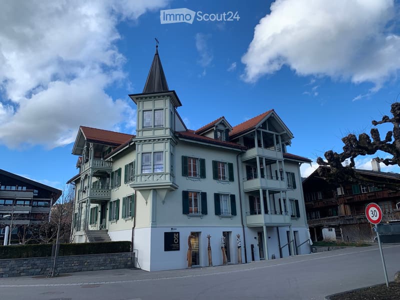 A three-story building with light green walls, red-tiled roof, and multiple balconies. Features a tower with a pointed roof and spire, green shutters, and decorative wooden railings.