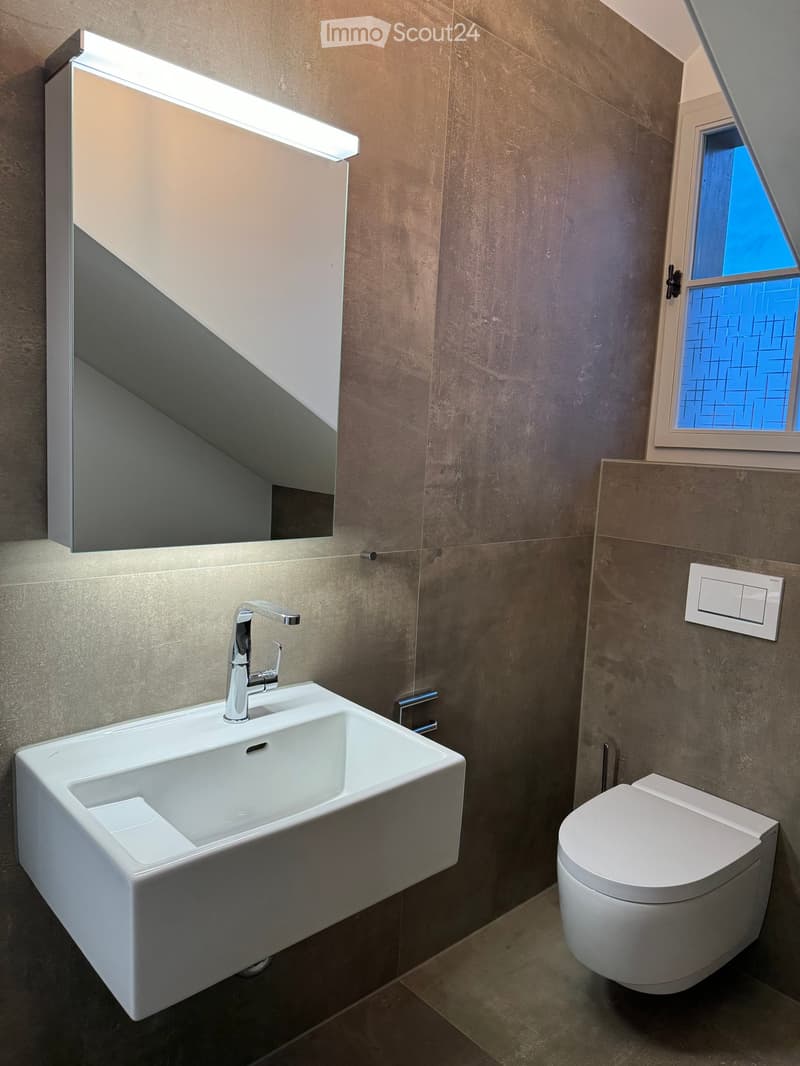 Modern bathroom with a white sink, chrome faucet, toilet, and large mirror with built-in lighting. The walls are covered in textured gray tiles, and there is a small window with blue-tinted glass.