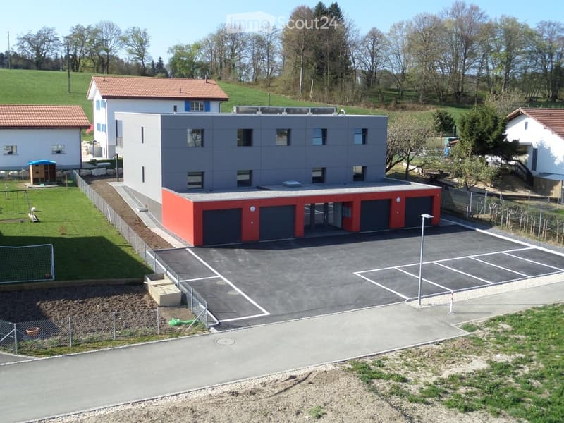 Modern two-story house with grey facade, red garage section, and large parking area.
