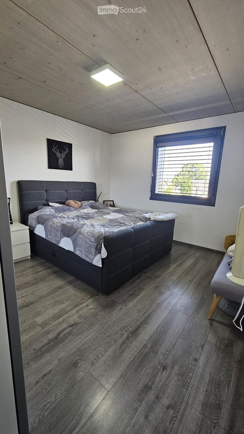 Modern bedroom with a large bed, nightstand, and a small stool with a lamp. The room features wooden flooring and a large window with blinds.