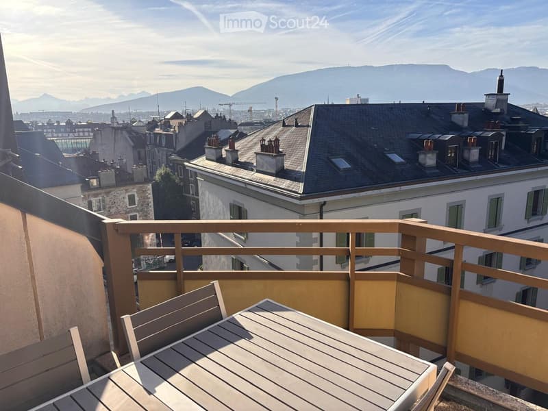 Balcony with wooden railing, table, and chair, overlooking cityscape and mountains.