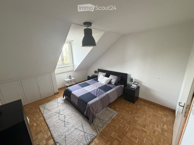 Modern attic bedroom with wooden flooring, large window, bed with grey and blue bedding, nightstands, and a hanging pendant light.