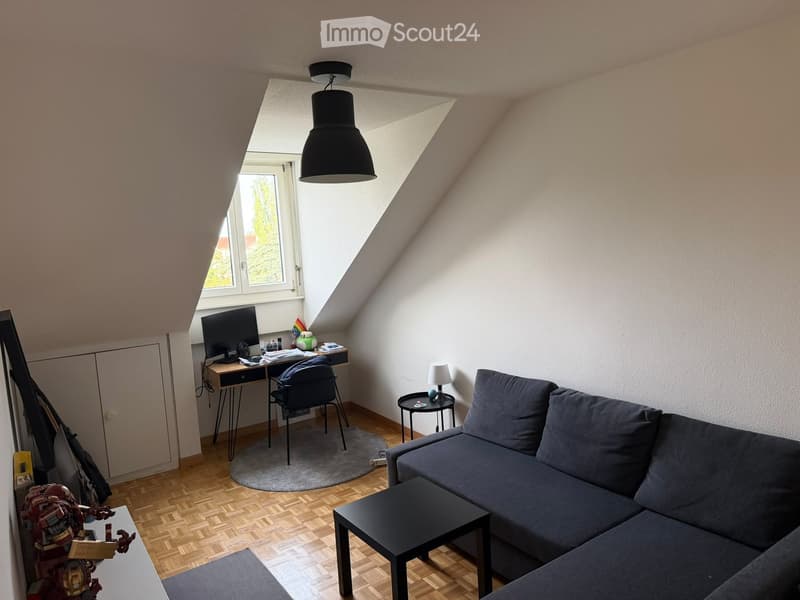 A cozy attic room with wooden flooring, a grey sofa, a coffee table, a desk with a computer, and a hanging pendant light.