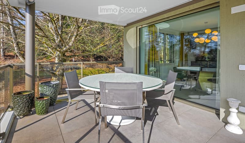Modern balcony with a round glass table and four grey chairs, surrounded by large green planters and a view of a garden.