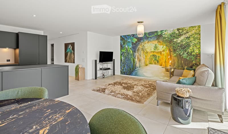 Modern living room with a large wall mural, a grey sofa with colorful cushions, a coffee table, a round dining table, and a TV stand with a flat-screen TV. The room features a sleek kitchen area with dark cabinets and a countertop.
