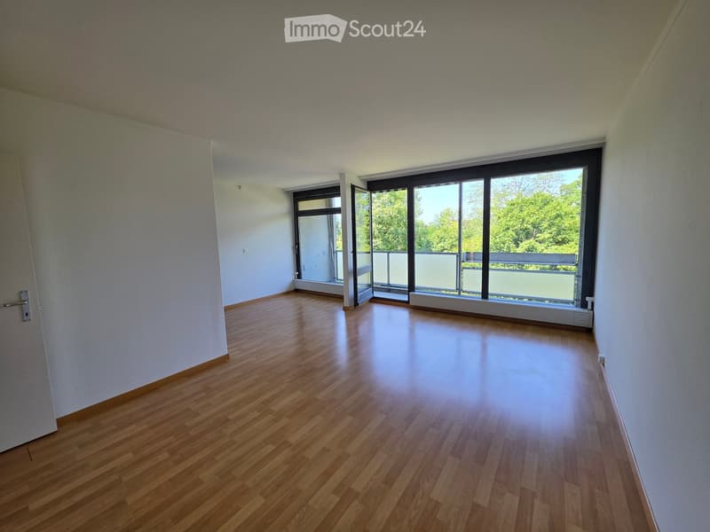 Empty room with wooden flooring, white walls, large windows, and a sliding door leading to a balcony.