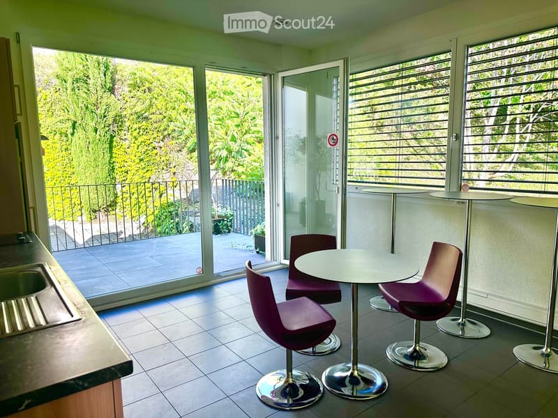 Modern interior with a round table and chairs, large windows and sliding doors leading to a balcony with greenery visible outside.