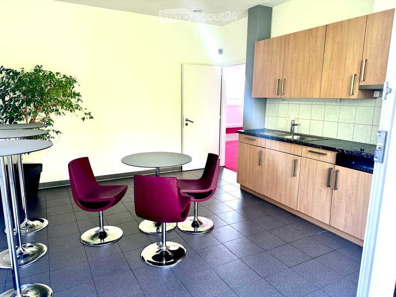 Modern, minimalist room with a small seating area featuring purple stool chairs and a round table, connected to a kitchen area with wooden cabinets, a sink, and a dishwasher.