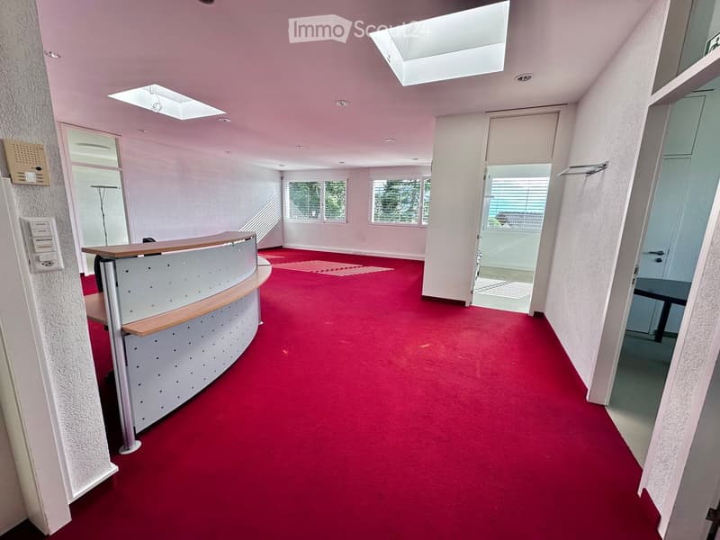 Open office space with red carpeting, a curved reception desk, and large windows with blinds. The area includes a door leading outside and a coat rack near the entrance.