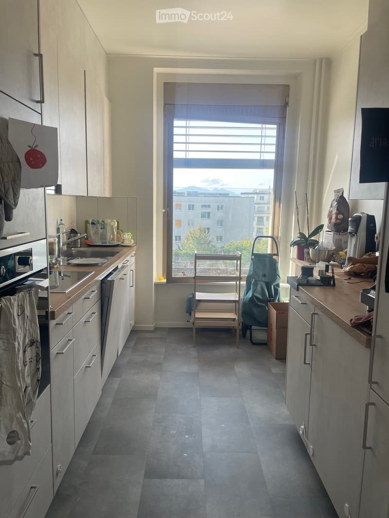 Modern kitchen with grey tiled floor, white cabinets, and stainless steel appliances. Features a sink, oven, dishwasher, refrigerator, microwave, coffee maker, and various kitchen items on the counter. A small table and chair are near the window, with a v
