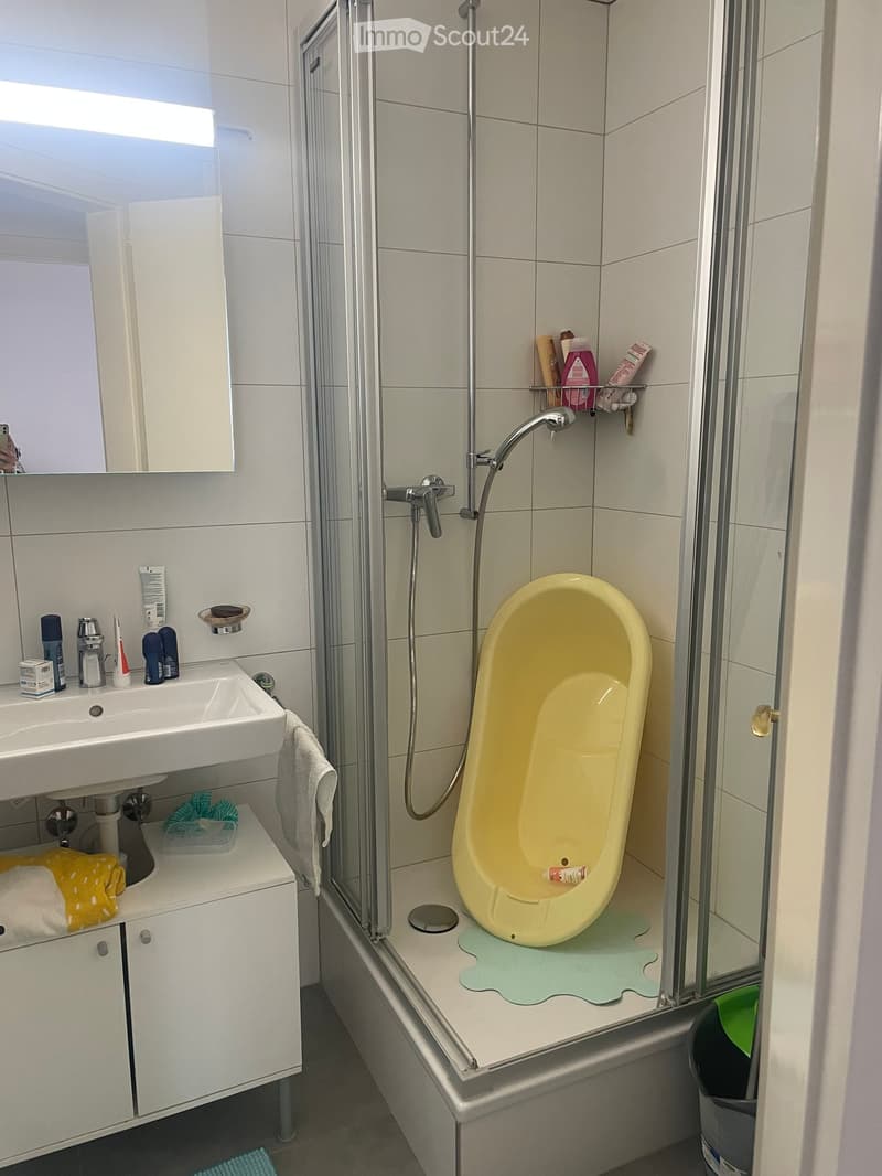 Modern bathroom with a white sink, mirror, shower, and a yellow toilet. The shower area has a glass enclosure and a modern faucet. The sink area includes a cabinet with towels and toiletries.