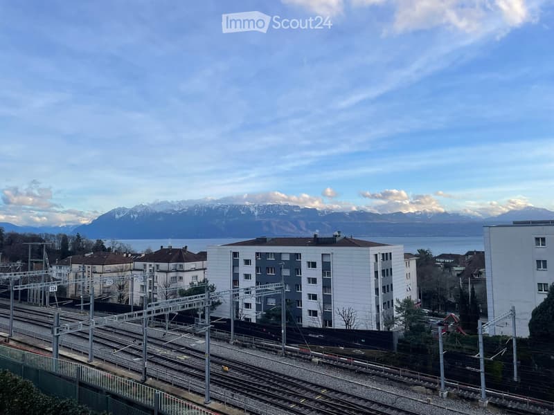 Modern multi-story residential building with white and grey facade, located near train tracks with a scenic mountain and lake view in the background.