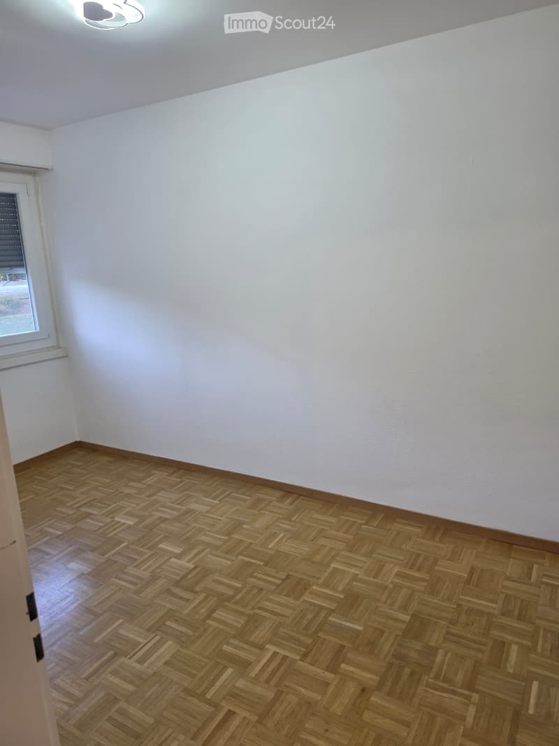 Empty room with wooden herringbone flooring, white walls, and a single window with blinds.