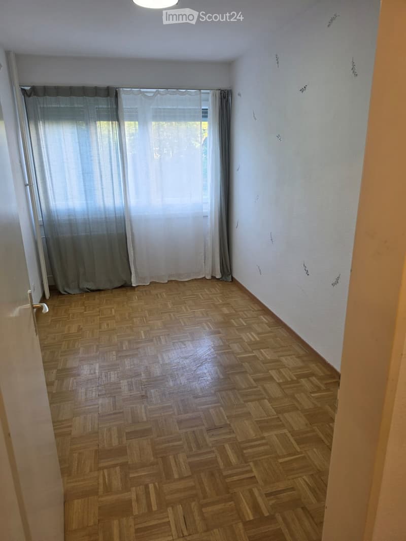 Empty room with wooden herringbone flooring, white walls, and large window with curtains.