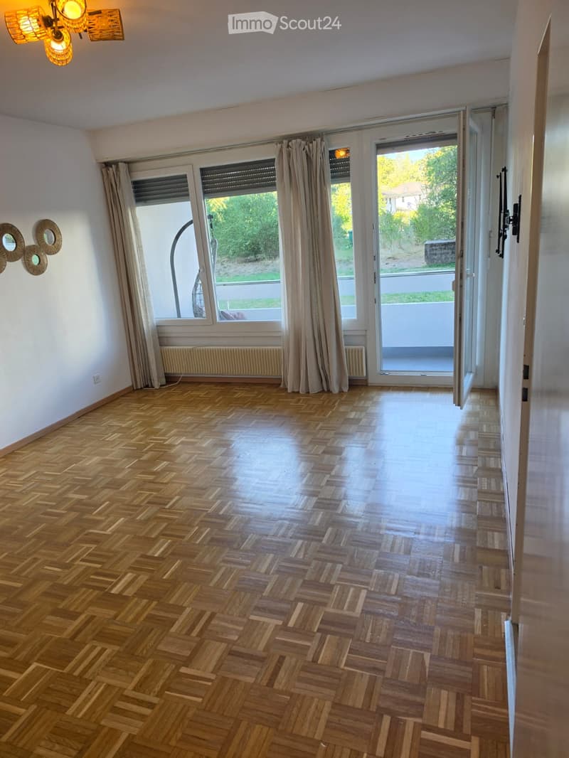 Empty room with wooden herringbone flooring, large windows with curtains, and a sliding glass door leading to a balcony.