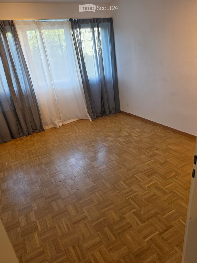 Empty room with wooden parquet flooring, white walls, and large windows covered with sheer and dark curtains.