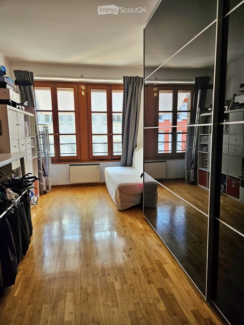 A modern, well-lit bedroom with wooden flooring, large windows, a single bed, a wardrobe, and a mirror. The room features a radiator and curtains, with a clothing rack visible on the left side.