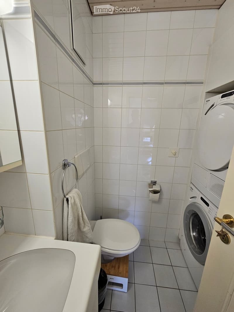 A compact bathroom with white tiled walls and floor, featuring a toilet, bathtub, washing machine, and a towel hanging on a ring.